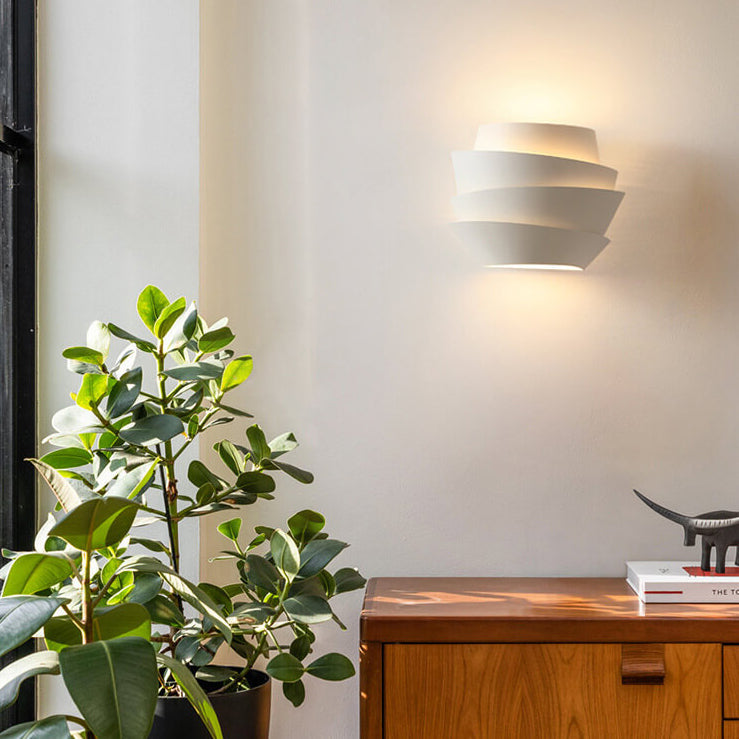 Moderne weiße Wandleuchte, geometrisches Design, neben Holzkommode und Zimmerpflanze.