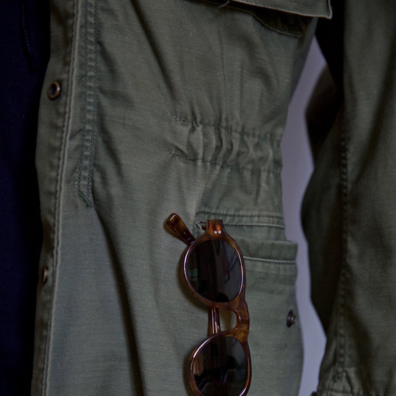 Nahaufnahme der unteren Taschenpartie einer grünen Herren Feldjacke mit praktischer Sonnenbrille eingesteckt.