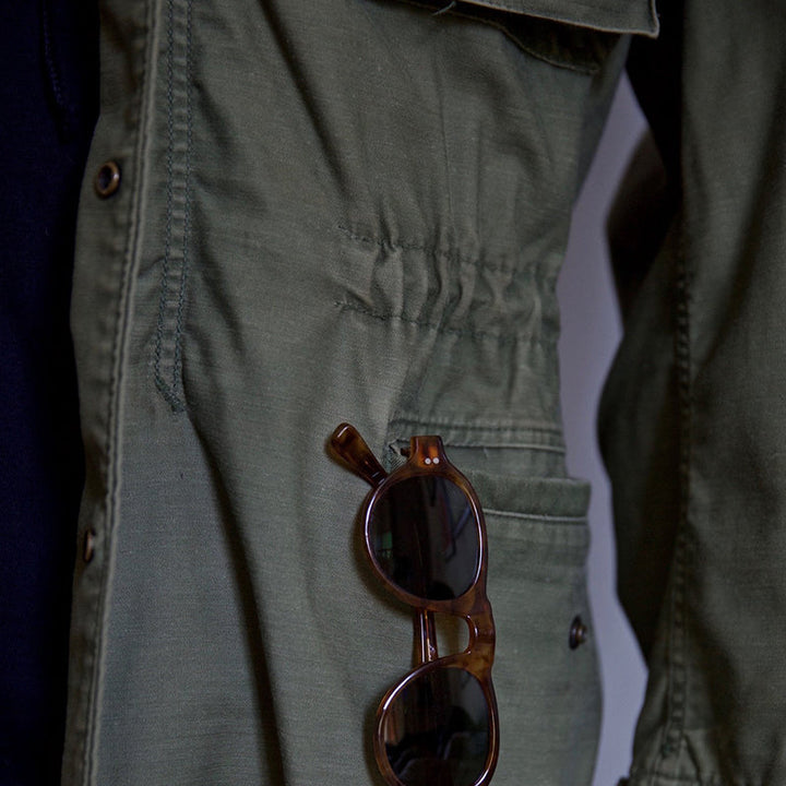 Nahaufnahme der unteren Taschenpartie einer grünen Herren Feldjacke mit praktischer Sonnenbrille eingesteckt.
