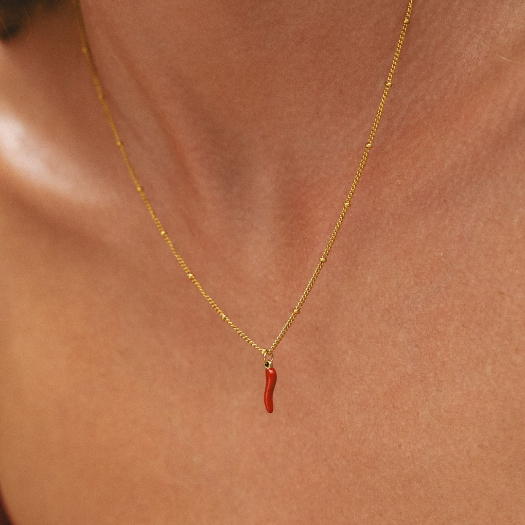 Nahaufnahme einer Frau mit goldener Halskette und rotem Chili-Anhänger am Dekolleté.