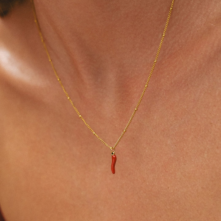 Nahaufnahme einer Frau mit goldener Halskette und rotem Chili-Anhänger am Dekolleté.